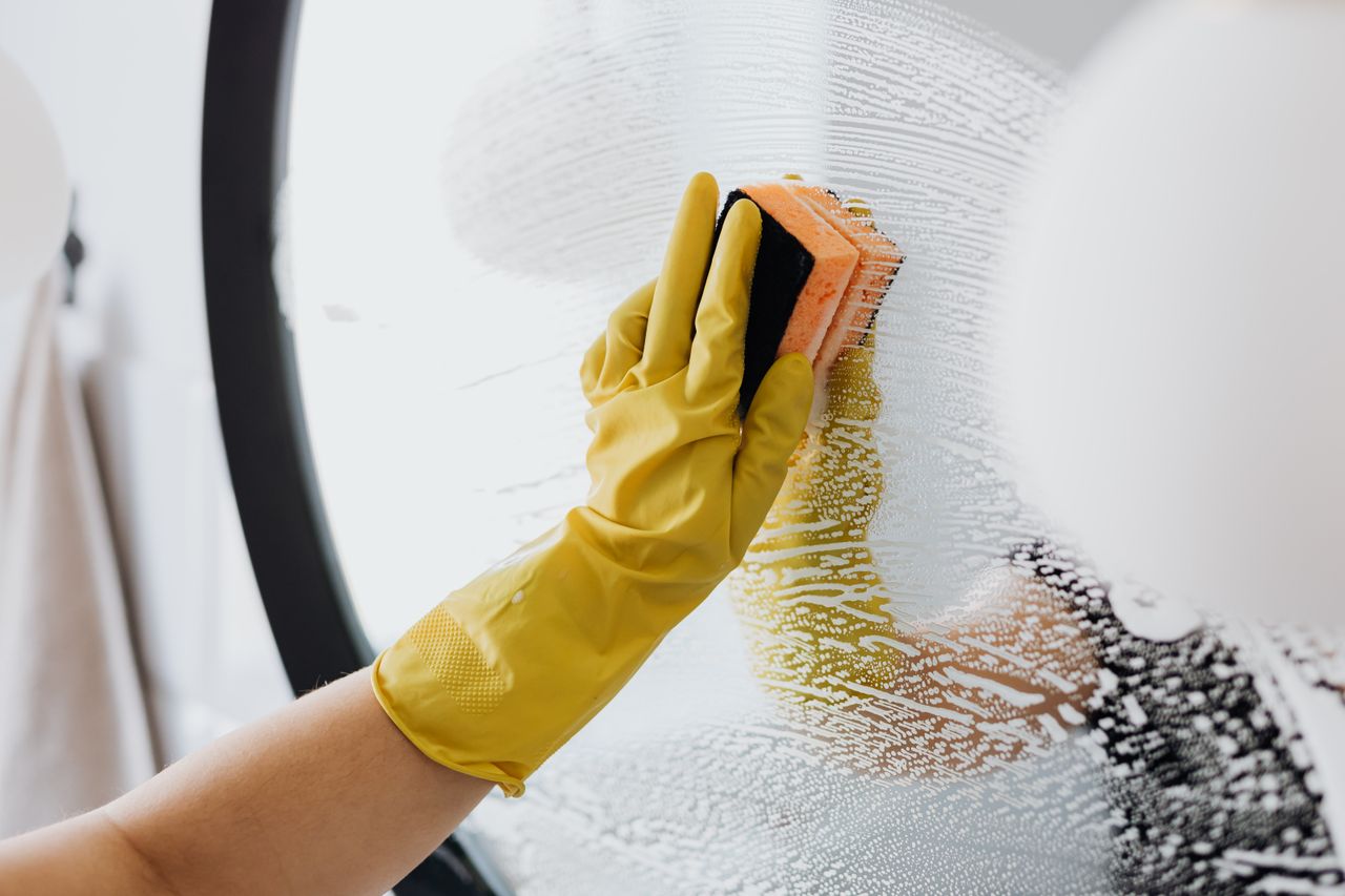 heros-img Personne anonyme portant des gants en caoutchouc jaunes et utilisant une simple éponge pour nettoyer le miroir dans la salle de bain